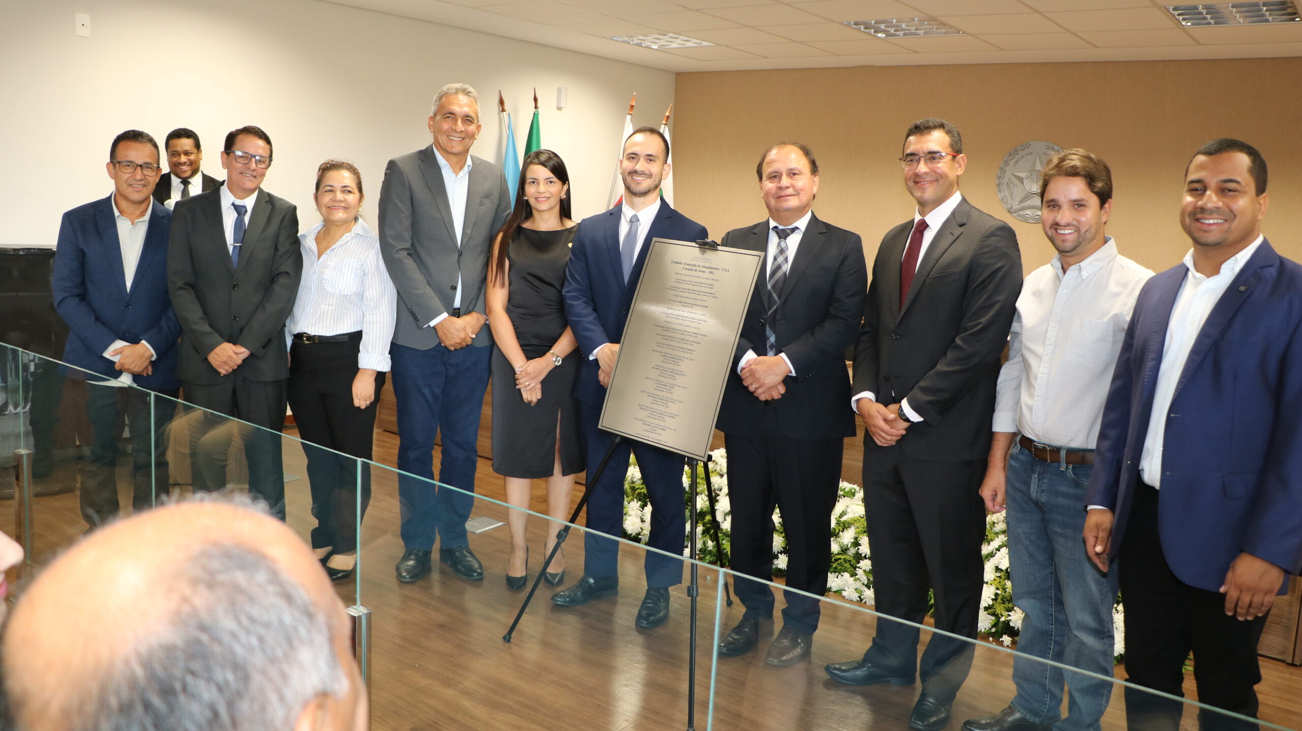 Grupo de pessoas posa para foto oficial em ambiente institucional, reunidas ao redor de uma placa comemorativa sobre um tripé. Os homens vestem ternos ou camisas sociais e a mulher usa um vestido preto, todos sorrindo diante de bandeiras ao fundo.