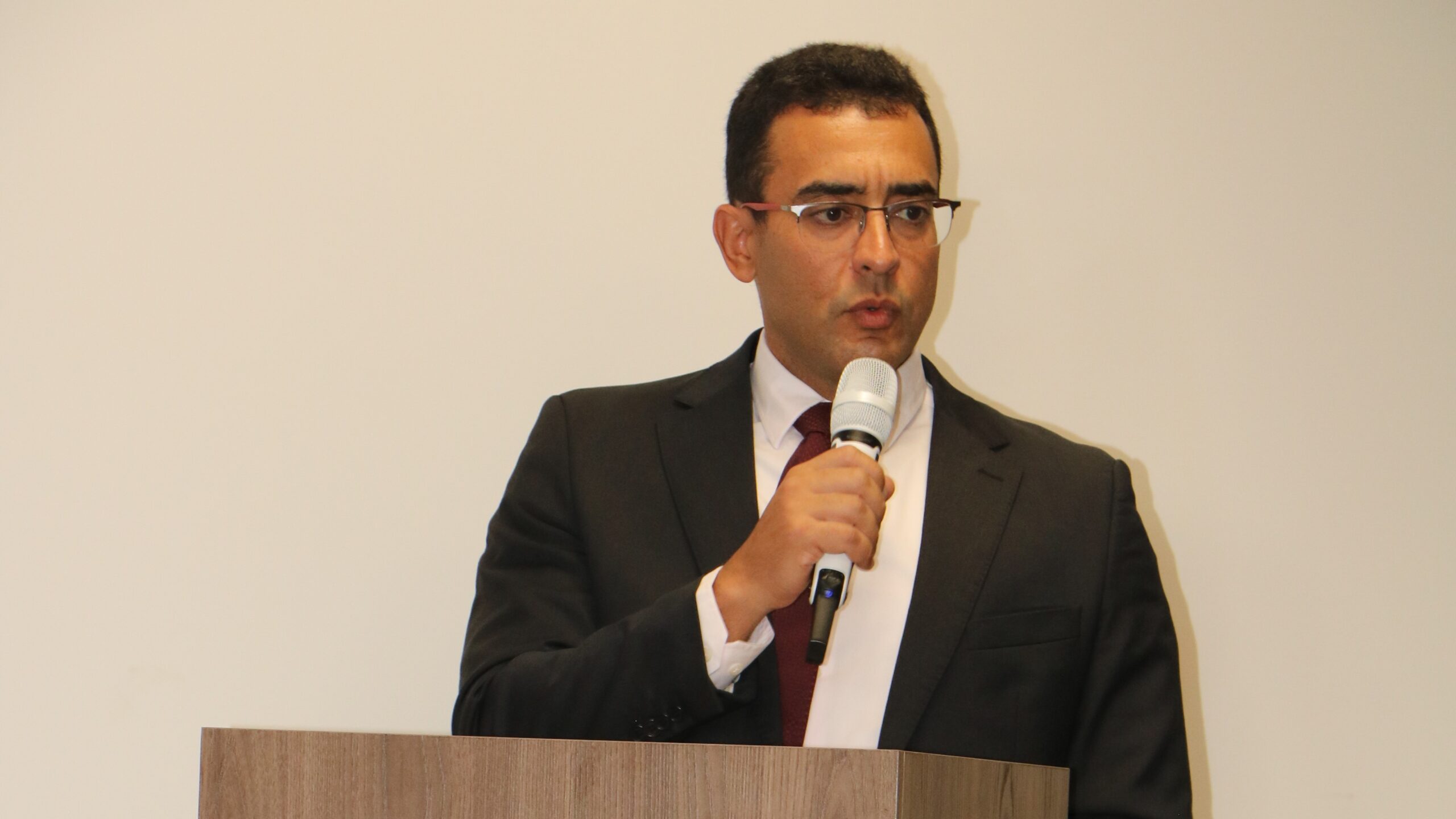 Homem de óculos, terno escuro e gravata bordô fala ao microfone em um púlpito de madeira. Ele está centralizado contra um fundo branco, em uma postura formal de discurso.