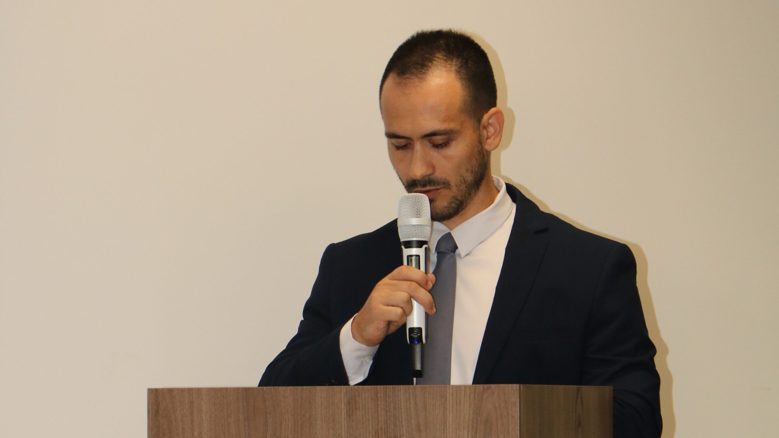 O homem de terno azul escuro e barba curta fala ao microfone em um púlpito de madeira, mantendo o olhar baixo em direção às suas notas. Ele está centralizado contra um fundo branco, em uma composição formal de discurso institucional.
