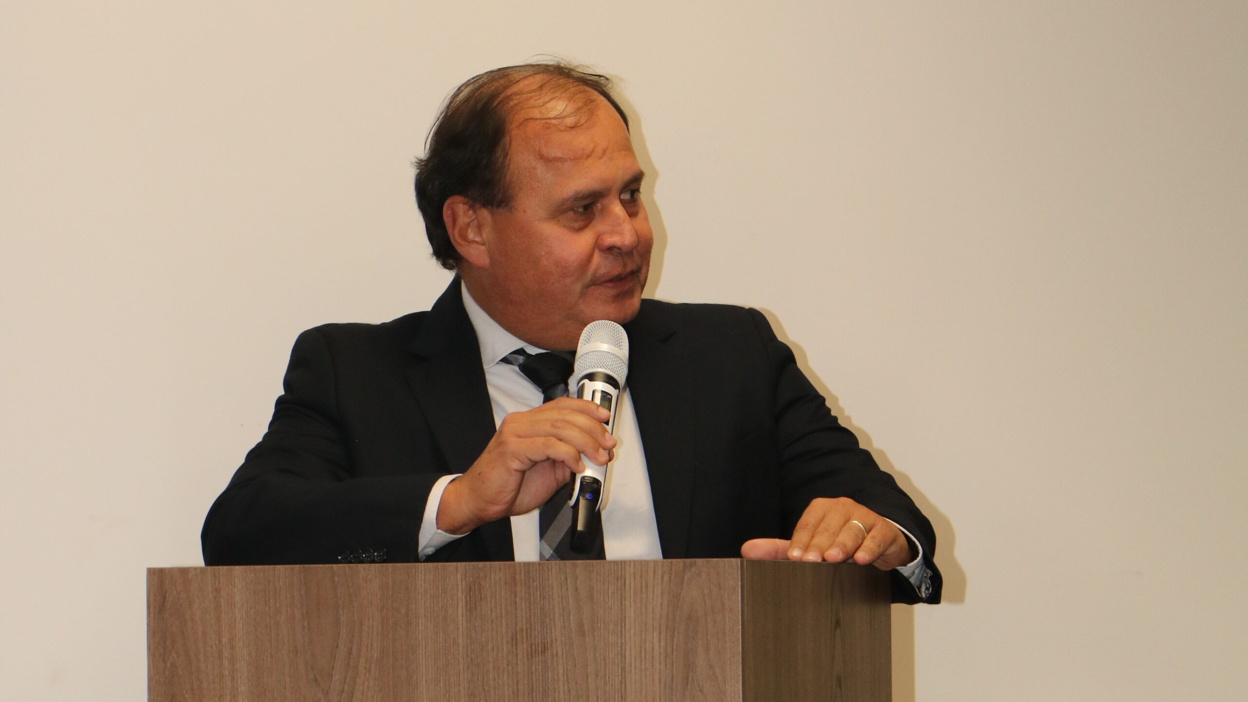 Homem de meia-idade, vestindo terno e gravata escuros, fala ao microfone em um púlpito de madeira. Ele está posicionado contra um fundo branco neutro, com o olhar direcionado levemente para o lado durante seu discurso.